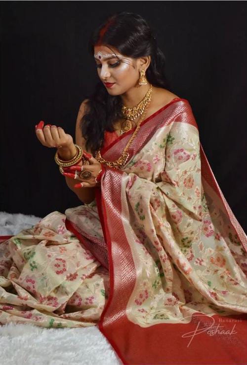 Woman in a floral saree with red border sitting on a white surface against a black background