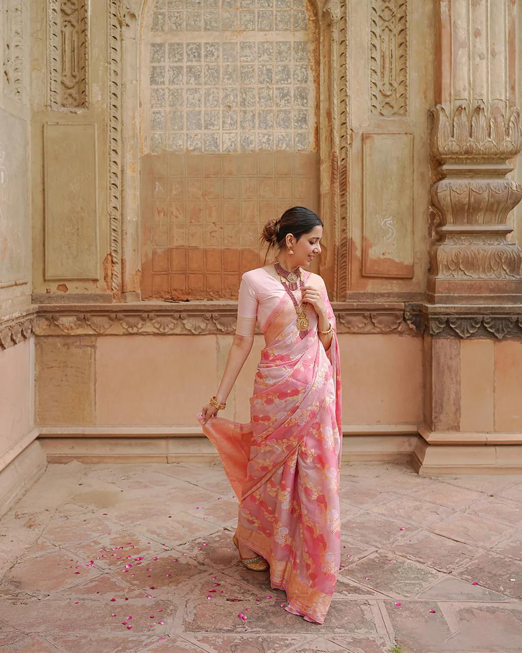 Woman in a pink saree standing in an ornate room with intricate wall designs.