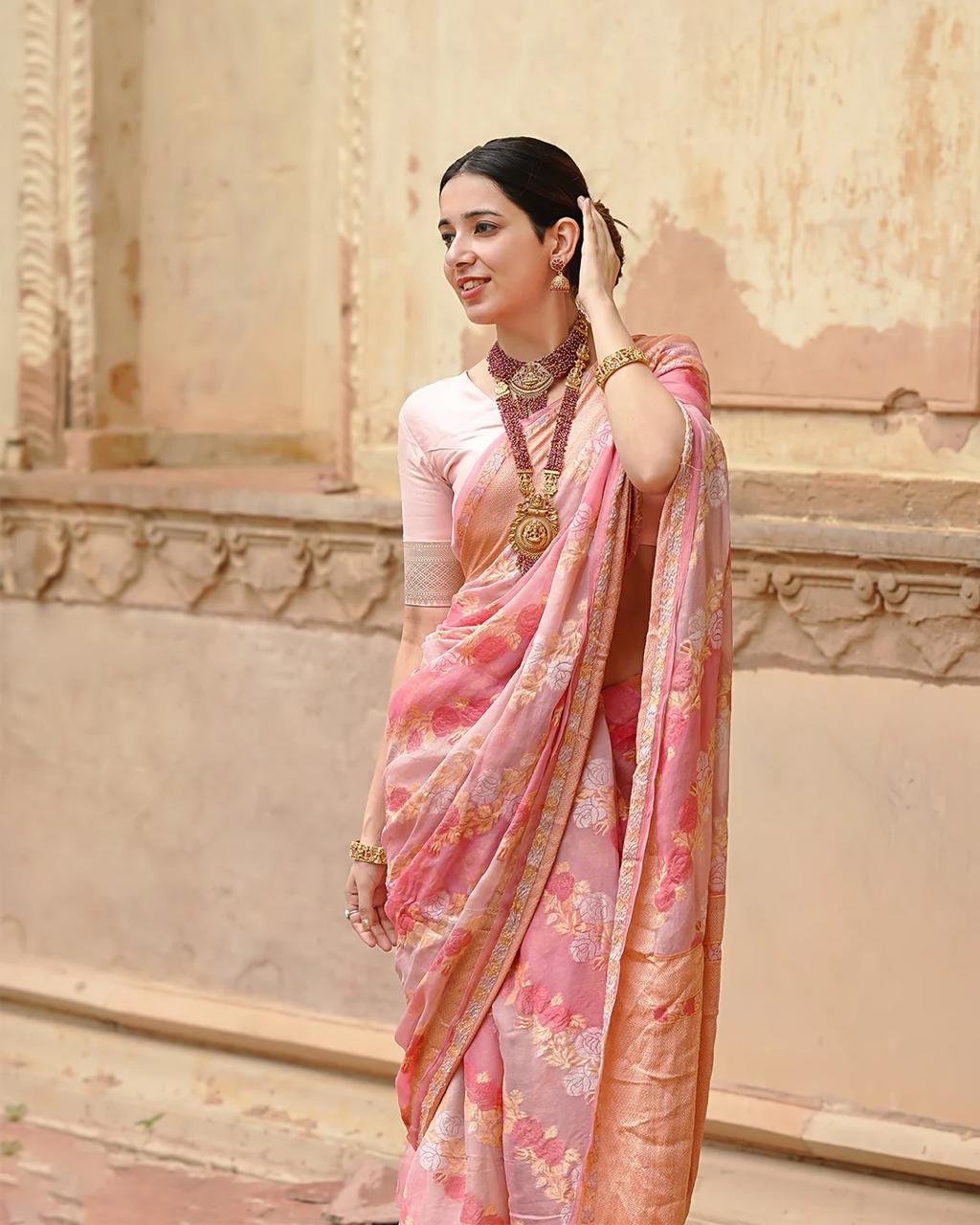 Woman in a pink saree with floral patterns standing against an architectural background.