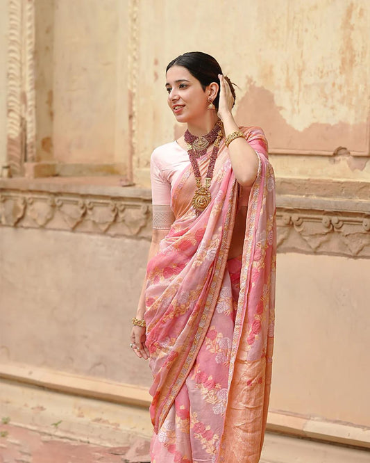 Woman in a pink saree with floral patterns standing against an architectural background.