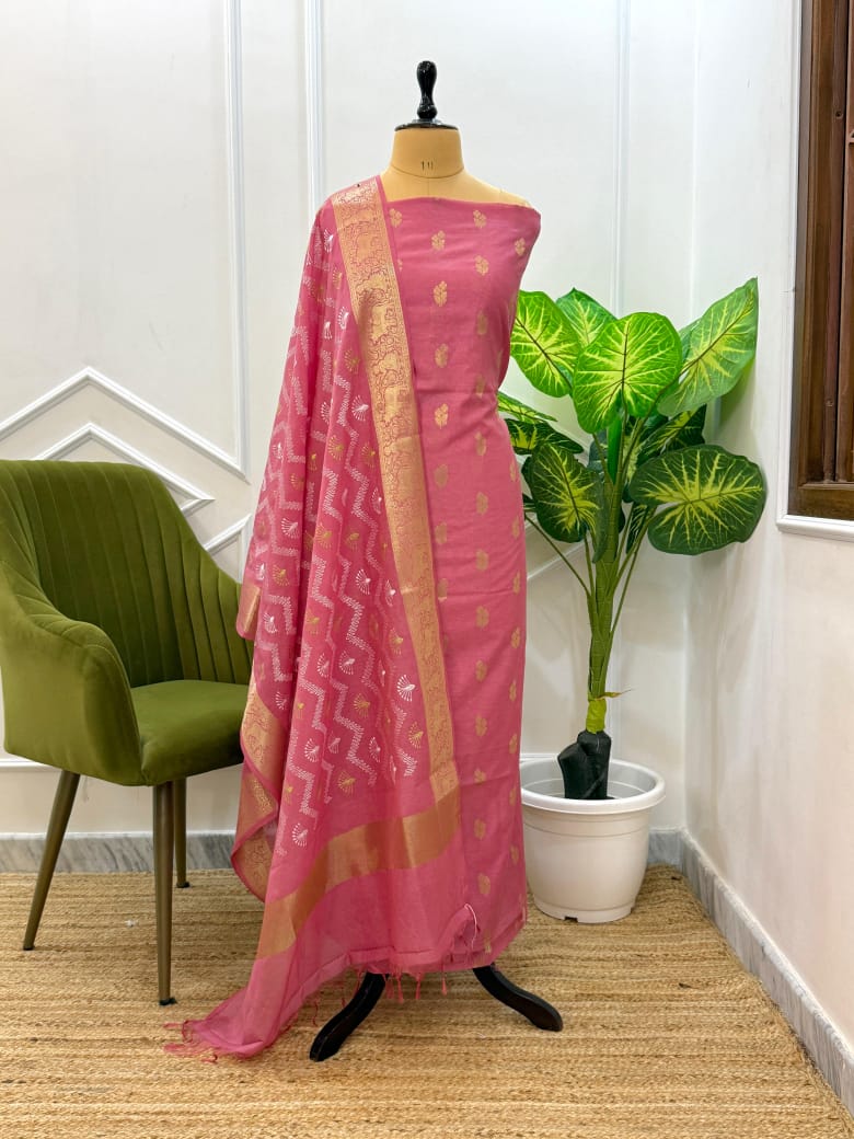 Pink saree with gold border on a mannequin against a white wall with a plant and chair.