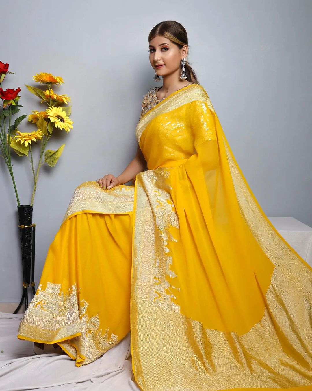 Woman wearing a yellow saree with a white blouse, sitting on a bed with flowers in the background.
