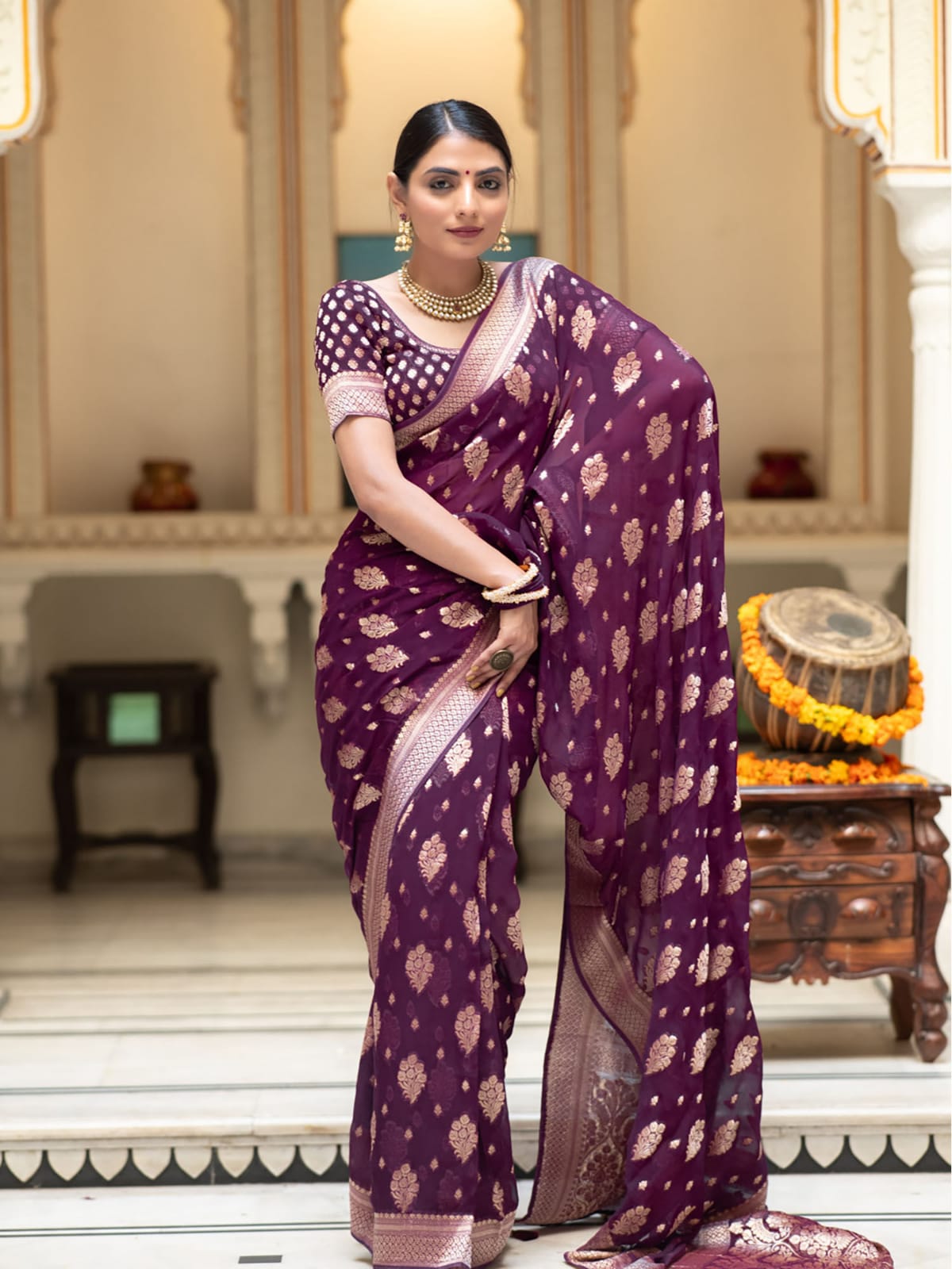 Woman in a purple saree with gold patterns standing indoors.