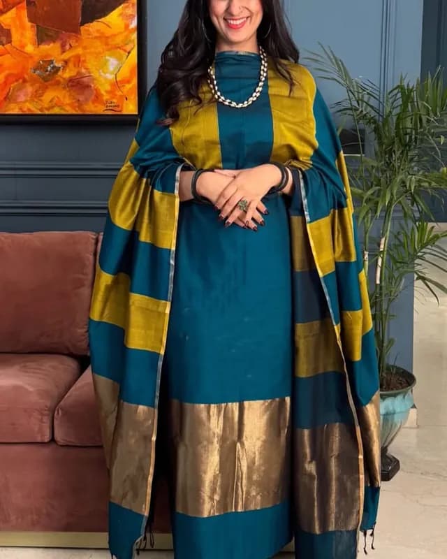 Woman in a teal and gold traditional outfit with a striped shawl, standing indoors.